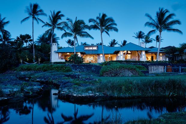 Night-time exterior view of a low, sprawling tropical residence with a blue metal hipped roof, pale rendered walls, illuminated windows and verandah-like openings, surrounded by tall palm trees and landscaped gardens beside a reflective waterway.