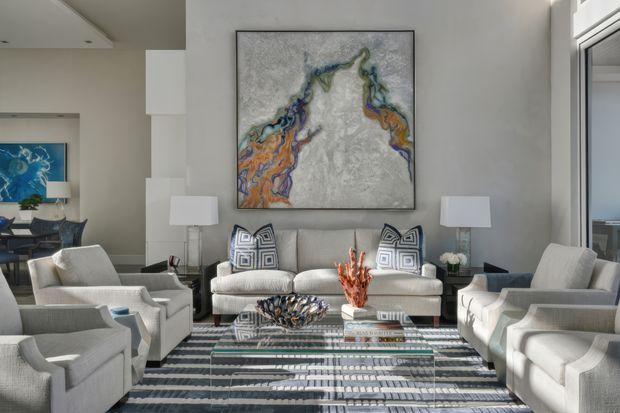 A bright, refined contemporary living room with a symmetrical seating arrangement of pale upholstered armchairs and a central sofa. A large abstract artwork forms the focal point above the sofa, complemented by geometric patterned cushions, glass side tables, sculptural decor, and a striped area rug. Floor-to-ceiling glazing is visible on the right, and an adjacent dining or lounge area appears in the background.