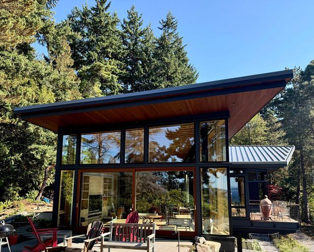 A modern two-storey glass-walled pavilion or house extension set among mature trees. The building features a dramatic low-pitched projecting roof with a warm timber soffit, dark-framed floor-to-ceiling glazing, timber-framed upper windows, and a metal-clad side roof. An outdoor terrace with casual seating is visible in the foreground and to the right, surrounded by a wooded landscape.