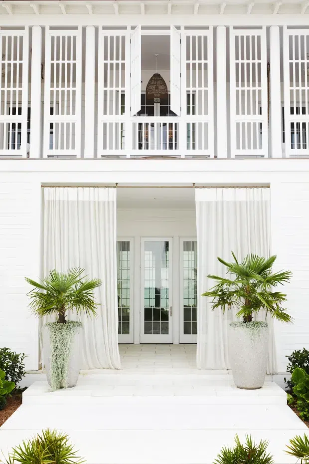 A bright white two-storey residential facade with a formal entry porch. The upper level features repeated tall louvered or gridded shutters, white columns, and a central open window with a hanging woven pendant visible inside. The lower entry has sheer white curtains, glazed French doors, white steps, and large textured white planters containing palm trees. The overall presentation is symmetrical, crisp, and tropical-coastal in character.