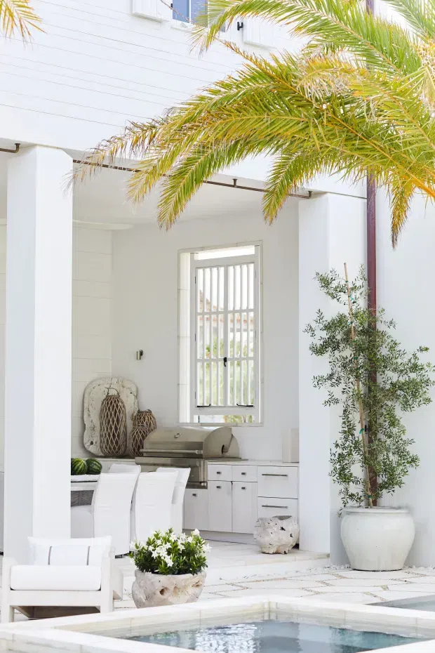 Bright white coastal-style outdoor entertaining area beside a swimming pool, with a covered outdoor kitchen and dining space framed by substantial white columns. The space features white built-in cabinetry, a stainless-steel barbecue, a barred window, tiled flooring, white seating, and tropical palms and potted greenery.