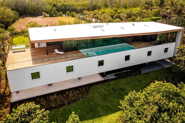 Aerial view of a long, contemporary white rectangular house set within dense tropical landscaping. The flat-roofed building features extensive dark glazing, a rooftop terrace with timber decking, and a long narrow swimming pool integrated into the upper-level outdoor space. Landscaped lawns, planting beds, and surrounding vegetation are visible around the structure.