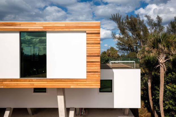 Exterior view of a contemporary two-storey house with a projecting upper volume clad in horizontal warm timber boards, crisp white rendered walls, large dark-framed glazing, and a partially visible rooftop terrace behind a glass balustrade. The building is elevated on angled supports and set among mature coastal-looking vegetation beneath a partly cloudy sky.