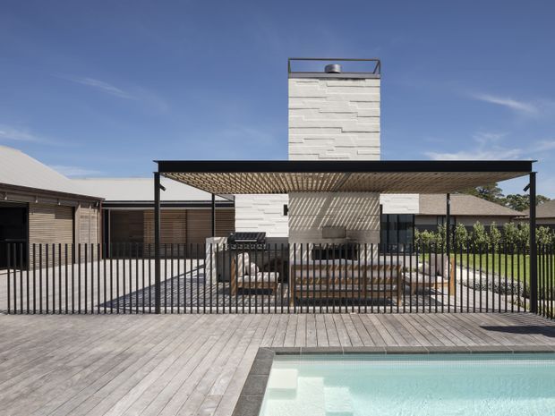 Contemporary single-storey residential exterior with a long flat-roofed pergola/carport-like structure, black metal posts and slatted overhead screening. A white textured masonry chimney tower rises behind it, with dark horizontal timber-clad walls and extensive timber decking surrounding a bright swimming pool. The foreground is enclosed by a vertical black metal fence, with neighbouring low roof forms visible at the sides.