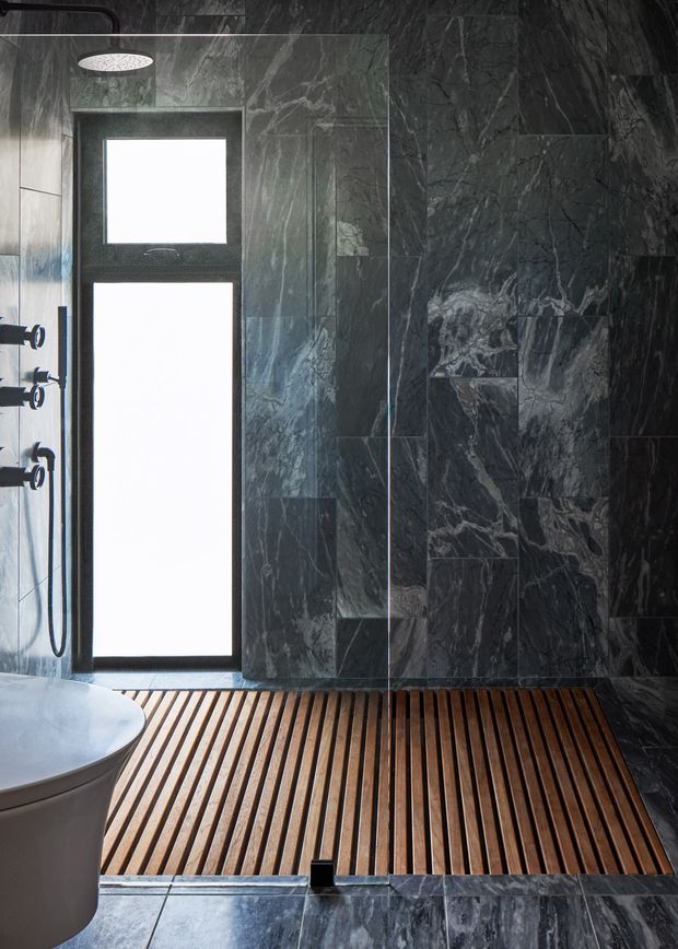 A dramatic, dark-toned contemporary bathroom featuring extensive grey-black veined stone wall and floor tiling, a frameless glass shower enclosure, wall-mounted shower fittings, a narrow vertical window, a white freestanding bathtub partially visible at left, and a warm slatted timber shower floor.