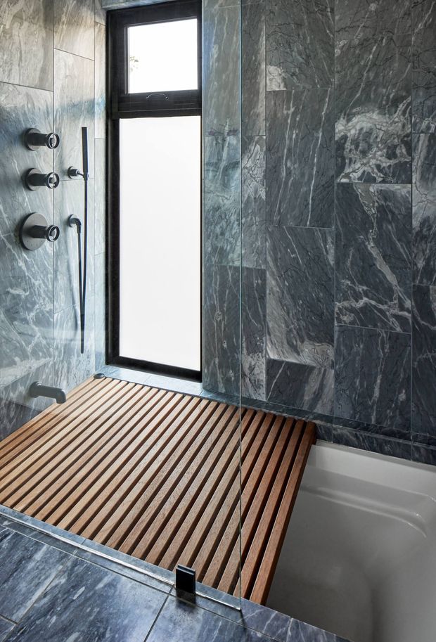 A contemporary bathroom featuring dark veined marble-look wall and floor tiles, matte black window and shower hardware, a frameless glass shower screen, and a warm timber slatted bath platform spanning the shower and bathtub. The space is compact, refined, and strongly material-led, with natural light entering through a tall vertical window.