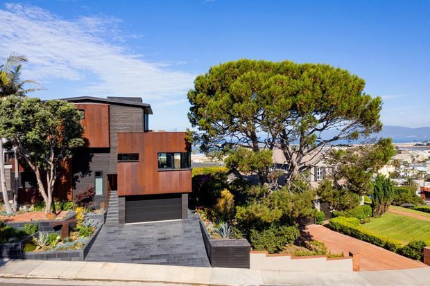 Exterior view of a contemporary multi-level residence on a sloping suburban site, featuring dark horizontal cladding, rust-toned metal-clad projecting volumes, a dark garage door, and a broad paved driveway. Landscaped garden terraces, mature trees, neighbouring homes, and a distant coastal or bay-like outlook are visible.