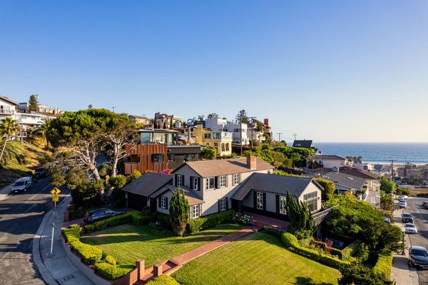 Aerial or elevated exterior view of a large residential home on a landscaped corner site in a dense coastal suburb. The dark painted house has multiple pitched gabled roof forms, contrasting timber-clad sections, white-trimmed windows, a broad manicured lawn, brick garden edging and paved paths. Other houses, streets and the ocean are visible in the background.