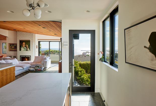 Bright open-plan residential interior viewed from a kitchen, with a large pale stone island in the foreground and a living room beyond. The living area includes a pale sectional sofa, fireplace, timber cabinetry, artwork, and a striking timber-lined angled ceiling. Full-height glazing and a glazed door open toward a hedge and ocean or coastal view. The space has white walls, dark-framed windows and doors, grey tiled flooring, and contemporary light fittings.