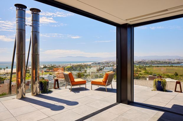 A bright contemporary terrace viewed through fully open corner glazing, with expansive tiled outdoor flooring, two orange lounge chairs and small side tables. Two tall polished metal flues or chimneys rise at the left, while the elevated outlook spans a coastal urban landscape, water, distant hills and cultivated fields. Dark-framed floor-to-ceiling glazing and a projecting roof create a strong indoor-outdoor connection.