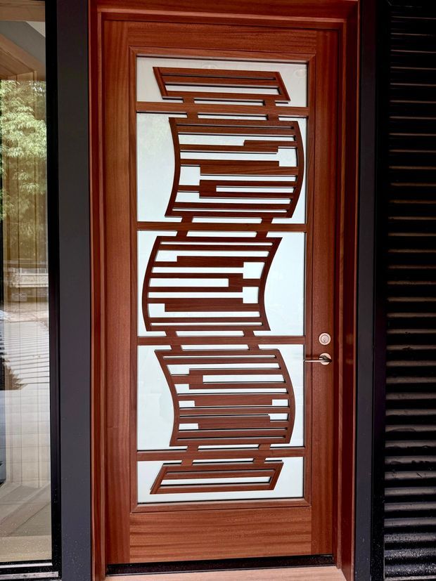 Close-up exterior view of a richly stained timber front door with a large decorative frosted-glass panel. The glass contains geometric, horizontally slatted timber or metal ornamentation arranged in curved bands, framed by substantial reddish-brown timber mouldings. A silver lever handle and lock are visible, with dark vertical exterior cladding or screening around the entry.