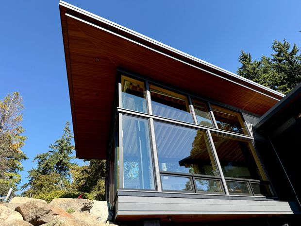 Exterior architectural photograph of a modern multi-level home set among trees and rock landscaping. The facade features extensive floor-to-ceiling glazing in dark metal frames, a strongly projecting low-pitched roof with a timber-lined soffit, and dark horizontal exterior cladding. The image is photographed from below against a clear blue sky.