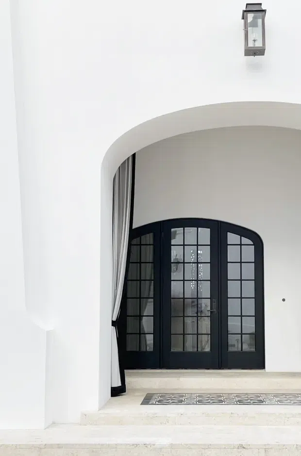 A bright, refined entryway framed by a large rounded arch. Pale stone or marble steps lead to a dramatic black multi-pane glazed double door, partly concealed by a light curtain. White plaster walls, a small dark wall lantern, and a patterned entry mat create a high-contrast, elegant composition.