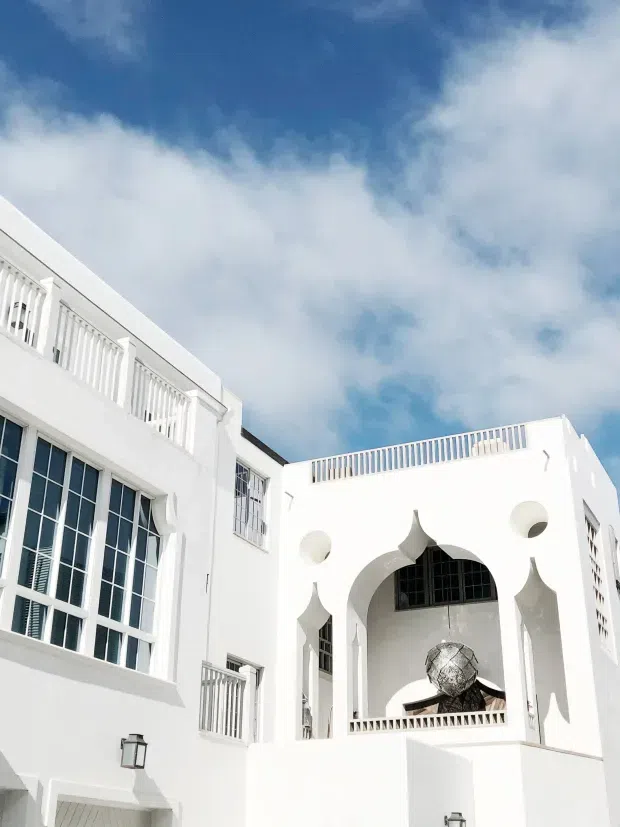 Bright white multi-level building exterior with Mediterranean or Moorish-inspired arched openings, geometric cutout details, decorative balustrades, large multi-pane windows, and a rooftop terrace. The facade is photographed from below against a partly cloudy blue sky.