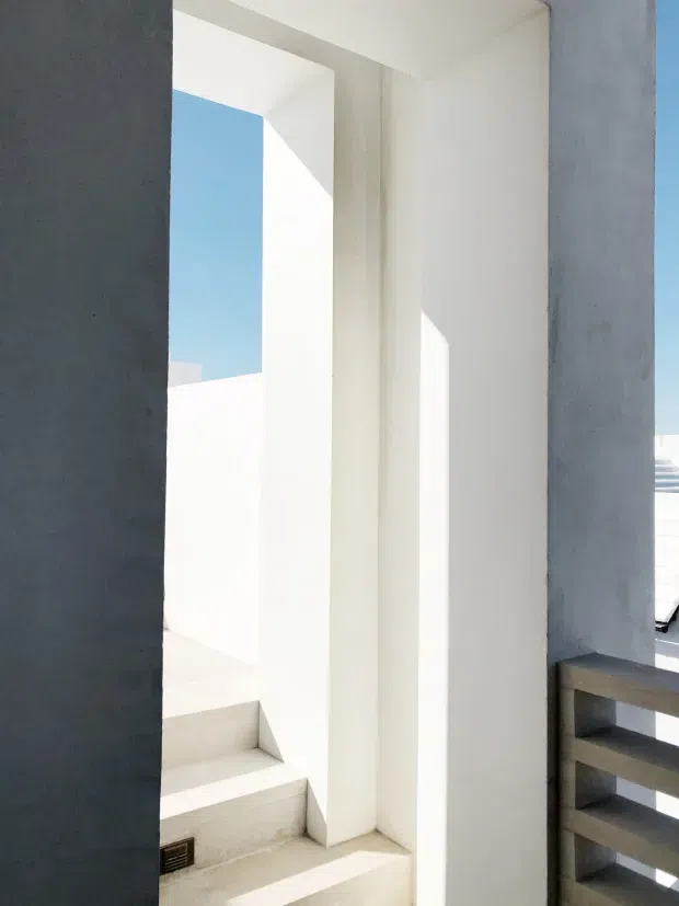 A bright minimalist architectural passage or stairway framed by stark white walls and deep rectangular openings. White concrete or plaster steps rise toward an opening with blue sky beyond, while a dark grey structural wall and grey horizontal balustrade contrast with the white surfaces. Strong sunlight creates crisp geometric shadows and highlights the building's clean, monolithic forms.