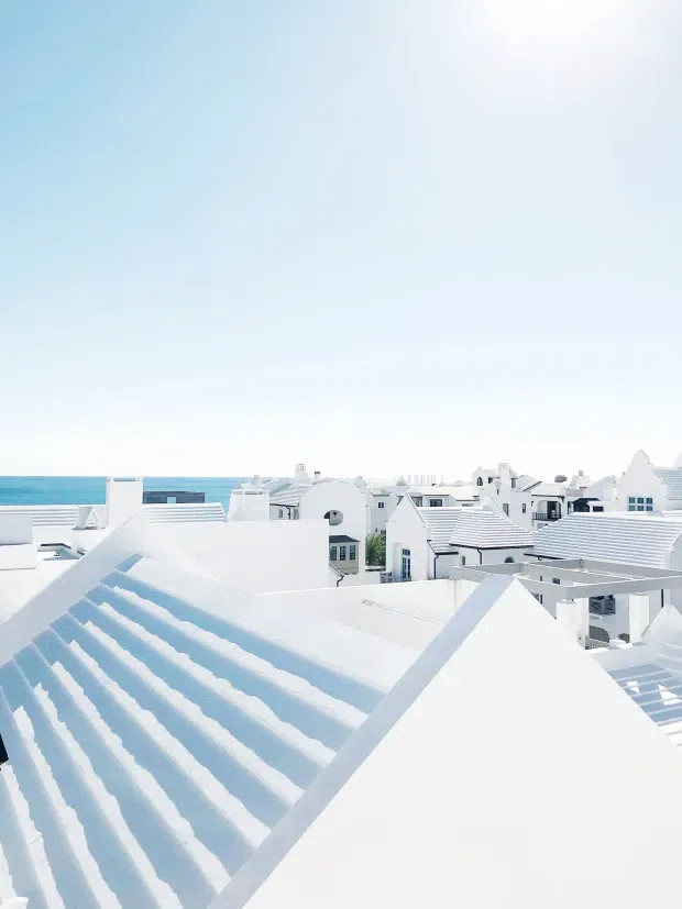 Bright elevated view across a dense coastal settlement of white buildings with repeated pitched and gabled roofs, white roof cladding, terraces, and parapets. The ocean is visible on the horizon beyond the rooftops. The image is dominated by the building roofs and surrounding seaside architecture under a pale blue sky.