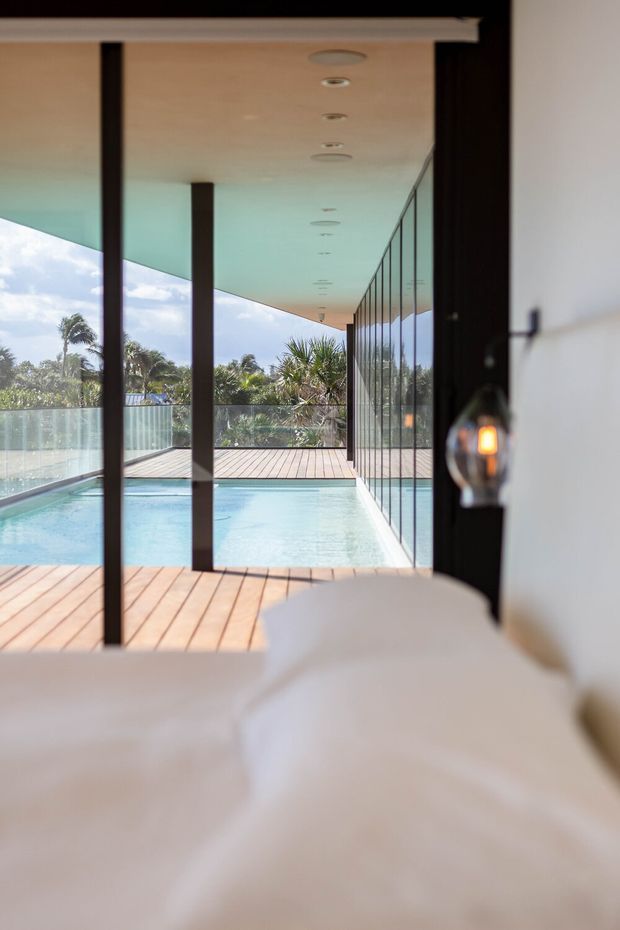 View from a bedroom toward a bright outdoor pool terrace, framed by extensive floor-to-ceiling glazing and dark slender structural posts. The room foreground contains a partially visible bed with white bedding and a wall-mounted pendant light; outside is a timber deck, swimming pool, glass balustrade and lush tropical planting. The architecture is clean-lined and contemporary with a deep projecting roof and recessed ceiling downlights.