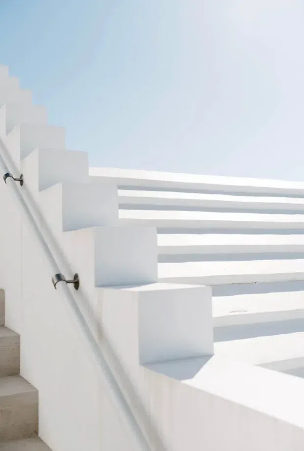 A bright, minimalist architectural detail showing a white stairway with solid stepped parapets, a slim metal handrail, and horizontal white balustrade elements casting crisp shadows. The scene is strongly sunlit with a pale blue sky visible beyond, creating a clean geometric composition.