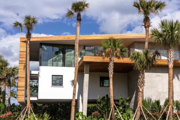 Exterior view of a contemporary two-storey residence with clean rectilinear massing, extensive dark-framed glazing, white rendered walls, exposed concrete columns, and warm timber soffits and cladding. The house is surrounded by landscaped tropical planting and palm trees under a partly cloudy sky.