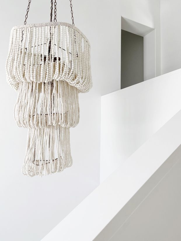 A close-up interior architectural detail featuring a large sculptural white beaded pendant chandelier suspended beside a crisp, minimalist white stair or balustrade. The space has smooth white walls, sharp geometric lines, and a recessed dark-grey opening in the background.