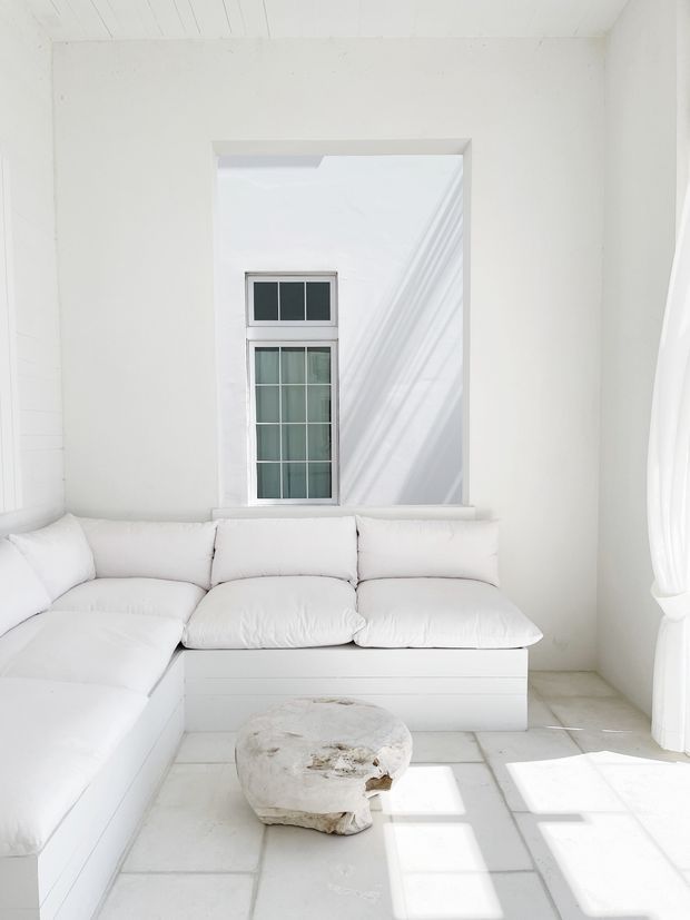 Bright, predominantly white living room with a low built-in sectional sofa beneath a tall multi-pane window. White tiled flooring, white walls and ceiling, sheer curtains, and strong sunlight create a highly minimal, airy interior. A rough organic stone or concrete coffee table provides the main contrasting feature.