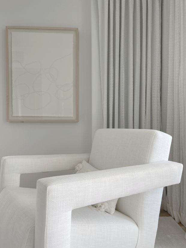 A close-up interior vignette featuring a sculptural white upholstered armchair positioned in front of full-height light grey sheer and draped curtains. A pale framed abstract artwork hangs on the soft grey wall, creating a monochromatic, refined and serene setting.