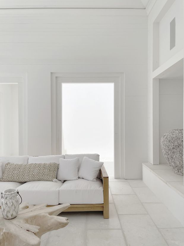 Bright, serene white-toned living room with a low timber-framed sofa, white cushions, a patterned neutral lumbar cushion, a rustic driftwood coffee table, and a decorative lantern. White horizontal wall panelling, a white tiled floor, deep window and opening trim, and a built-in display ledge create a clean coastal interior. A large bright window provides natural light, while a textured spherical object sits in the built-in recess.