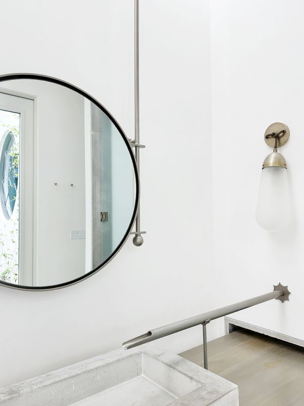 Bright minimalist interior detail featuring a white plaster wall, large round black-framed mirror, sculptural wall sconce, and a pale grey stone or concrete vanity basin. A slim metal handrail and stair or landing edge are visible in the foreground, with light timber flooring beyond.