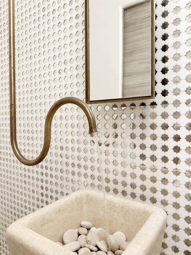 Close-up view of a compact bathroom basin with a sculptural aged-bronze wall-mounted faucet, flowing water, and a rectangular mirror. The walls are covered in a repeating white and light-grey quatrefoil mosaic tile pattern, while the basin is a pale textured stone with decorative smooth pebbles inside.