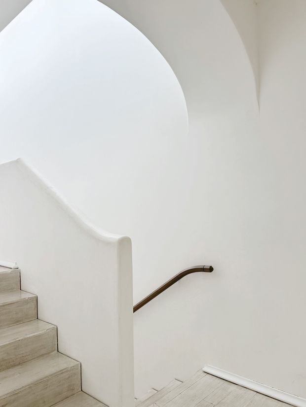 Minimalist interior view of a curved stairway with smooth white plaster walls and sculptural balustrade, pale timber or concrete-finished steps, and a dark bronze-brown handrail following the stair curve. The composition emphasizes the sweeping arch and clean architectural geometry.