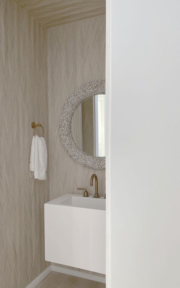 A compact, refined bathroom powder room viewed through a partially open doorway. The space features a wall-mounted white vanity with an integrated rectangular basin, brushed brass/gold tapware, a matching brass towel ring with white hand towels, pale textured leaf-pattern wall covering, and a large round mirror framed with a shell-like natural textured border. The palette is soft neutral and warm, with light flooring and white trim.