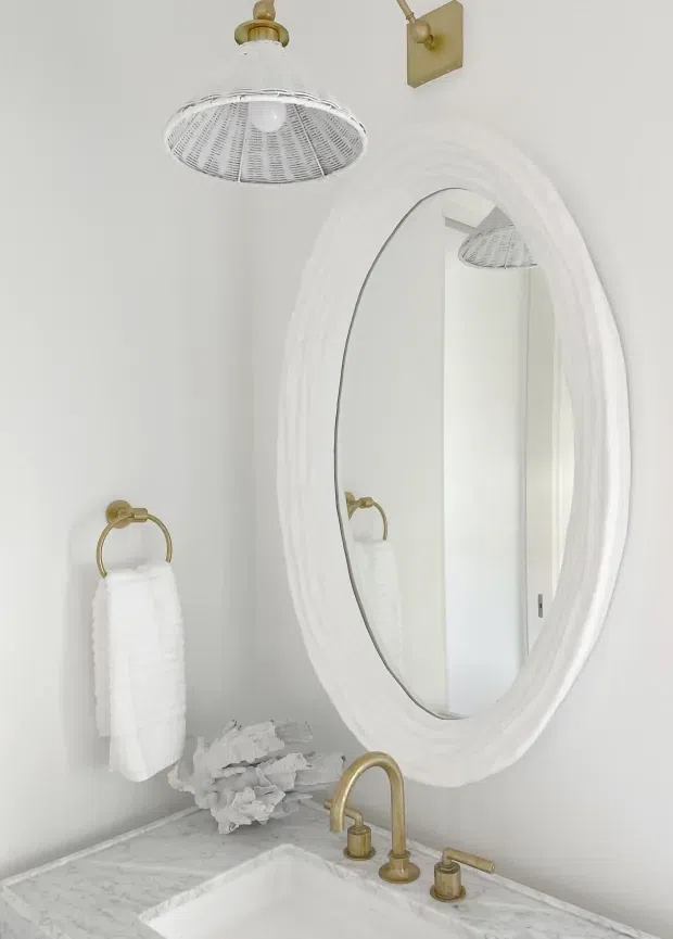 A bright, compact bathroom vanity area with a white oval framed mirror, marble countertop, white basin, and brushed brass tapware. A brass towel ring with a white hand towel, sculptural white decor, and a woven pendant light contribute to a soft, refined coastal-inspired look.