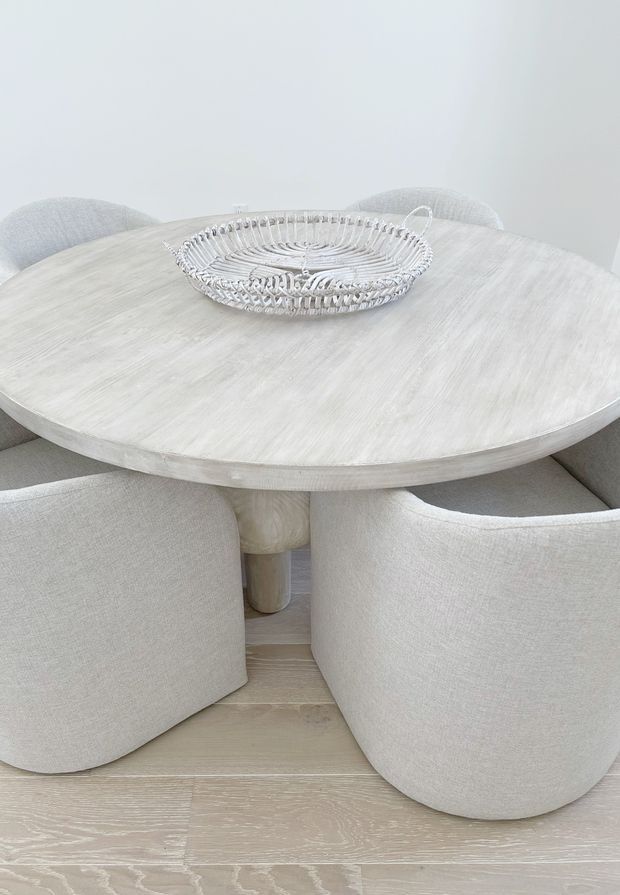 Bright, softly styled dining area featuring a round, whitewashed timber dining table with a central pedestal base, surrounded by curved upholstered dining chairs in pale neutral fabric. A delicate silver-toned wire basket sits on the tabletop. The room has pale timber flooring and a minimal, airy neutral palette.