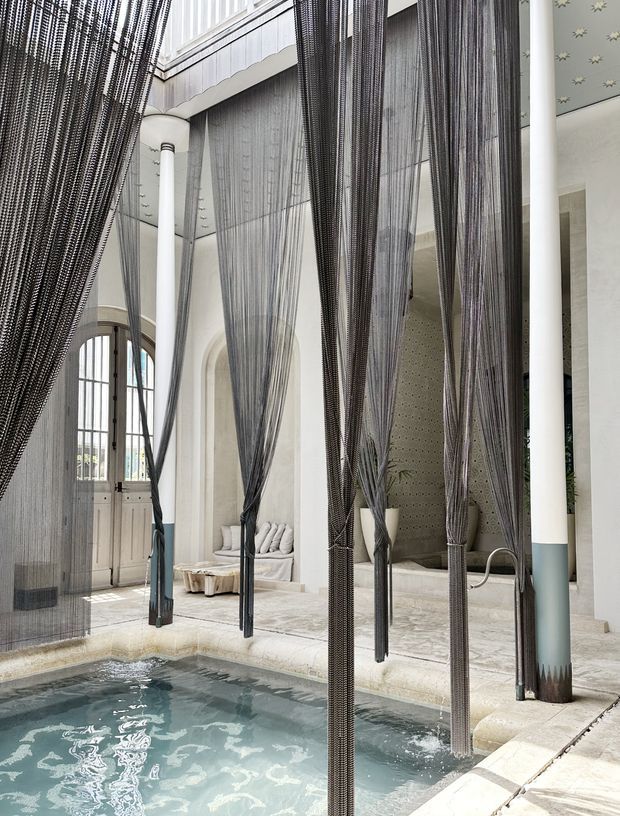 A refined indoor courtyard pool with pale stone paving, arched openings, slender white columns, and tall gauzy dark mesh curtains suspended around the water. A built-in cushioned seating nook and patterned ceiling add decorative detail, creating a serene, light-filled spa-like environment with a contemporary Mediterranean or Moroccan-inspired character.