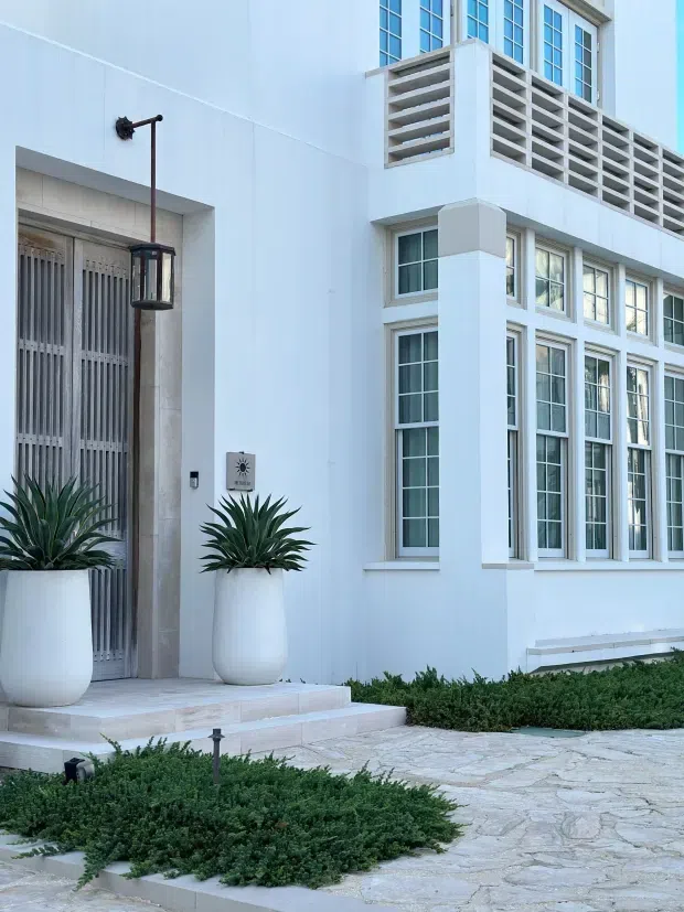 Exterior view of a refined white stucco building entrance with tall multi-pane windows, a decorative louvred parapet, a vertically slatted entry door, hanging lantern, and large white planters with agave. Pale natural-stone paving and clipped low hedges frame the façade.