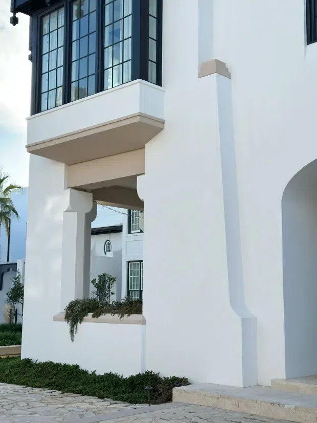 Close exterior view of a white, multi-storey house with strong symmetrical architectural detailing, tall black-framed divided-light windows, projecting balcony, decorative column-like supports, arched opening, and pale stone paving. Low landscaped planting borders the facade, with another similarly styled white building visible beyond.