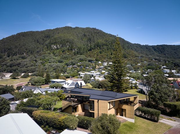 Aerial exterior view of a contemporary single-storey timber-clad house with a dark low-pitched roof, large windows and a partially sheltered outdoor terrace. The home sits in a landscaped suburban neighbourhood, with dense forested hills rising behind it.