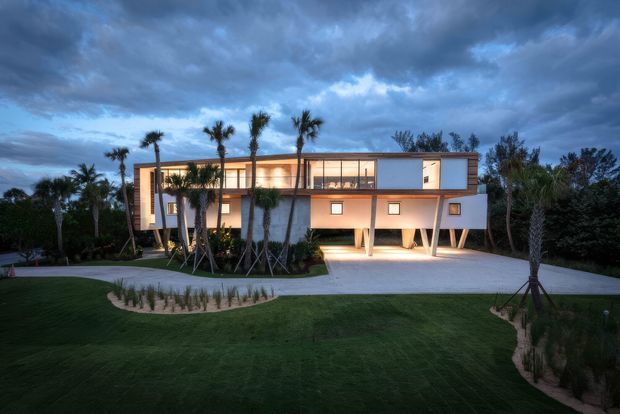 A contemporary two-storey residence photographed at dusk, set within a landscaped garden. The building has a long rectilinear form with extensive upper-level glazing, warm interior illumination, pale rendered wall surfaces, dark slim window frames, and a broad flat roof or very low-profile roofline. The lower level is partially elevated on angled supports, creating a sheltered open area and driveway beneath. Palm trees and structured planting surround the house.