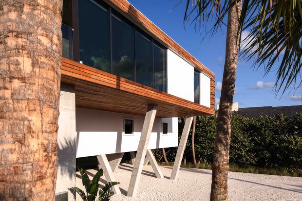 Exterior view of a contemporary elevated house in a tropical setting. The upper level projects outward with expansive dark glazing, warm timber soffits and cladding, white rendered walls, and angled structural supports beneath. Palm trees, sandy ground, and a bright blue sky contribute to the coastal atmosphere.