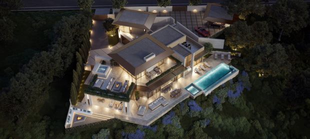 Aerial dusk rendering of a large multi-level residential property surrounded by dense landscaped vegetation. The home has several pitched roof volumes, extensive outdoor terraces, a long illuminated swimming pool, a spa or plunge pool, and warm architectural lighting.