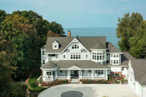 Aerial view of a large ornate light-coloured multi-storey house overlooking a blue body of water. The home has a complex grey shingle roof, multiple gables and dormers, projecting bay windows, a rounded turret-like porch, white decorative railings and trim, and a broad circular driveway surrounded by mature trees and landscaped gardens.