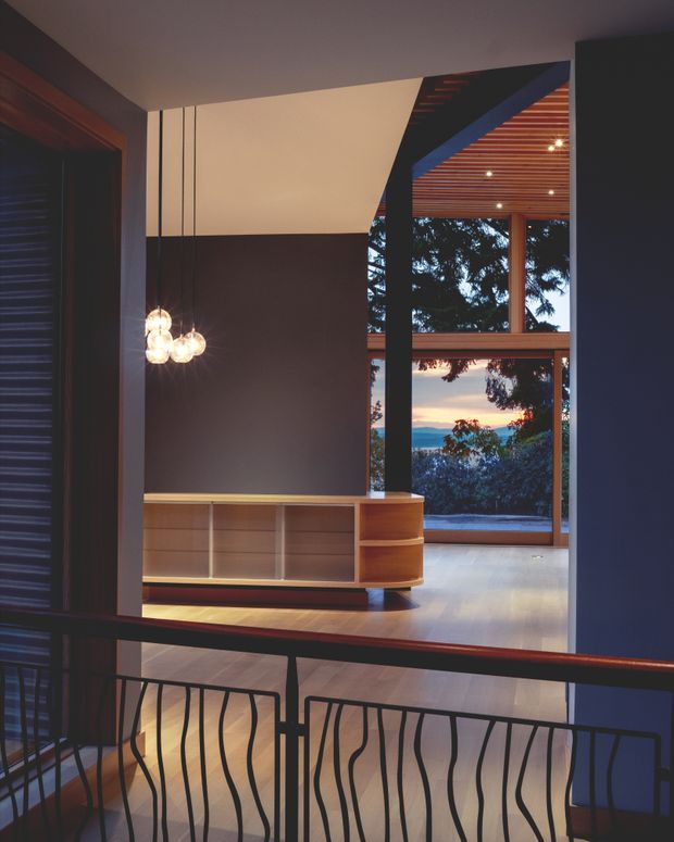 Interior view looking into a refined open-plan living area, with dark painted walls, warm timber flooring, a low built-in timber media/storage cabinet, and sculptural pendant lights. A dramatic black structural frame opens toward a glazed outdoor-facing wall with timber-framed windows, a slatted timber ceiling, and distant landscape views. A decorative dark metal balustrade occupies the foreground, suggesting an upper-level landing or stair edge. The space has a strong indoor-outdoor connection and a warm evening atmosphere.