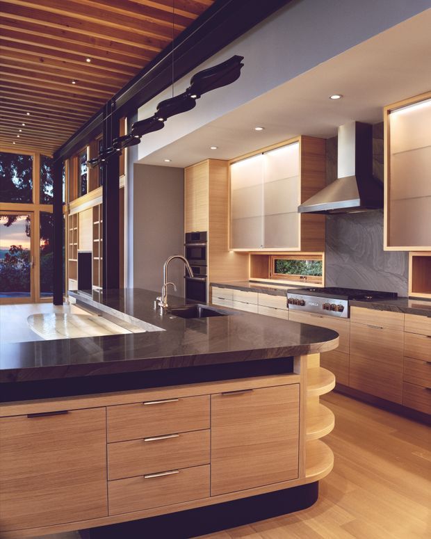 A warm, contemporary kitchen with extensive light timber cabinetry, a large dark stone island with curved edges and integrated seating shelves, stainless-steel appliances, and a textured dark splashback. The kitchen opens toward a tall glazed living area with dramatic exposed timber ceiling battens and dark structural beams. Under-cabinet and recessed lighting create a refined, inviting atmosphere.