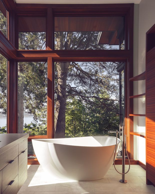Bright contemporary bathroom featuring a freestanding white bathtub positioned in front of expansive timber-framed windows. The room has warm dark timber wall panelling and shelving, pale large-format floor tiles, a minimalist freestanding bath filler, and abundant natural light with leafy trees visible outside.