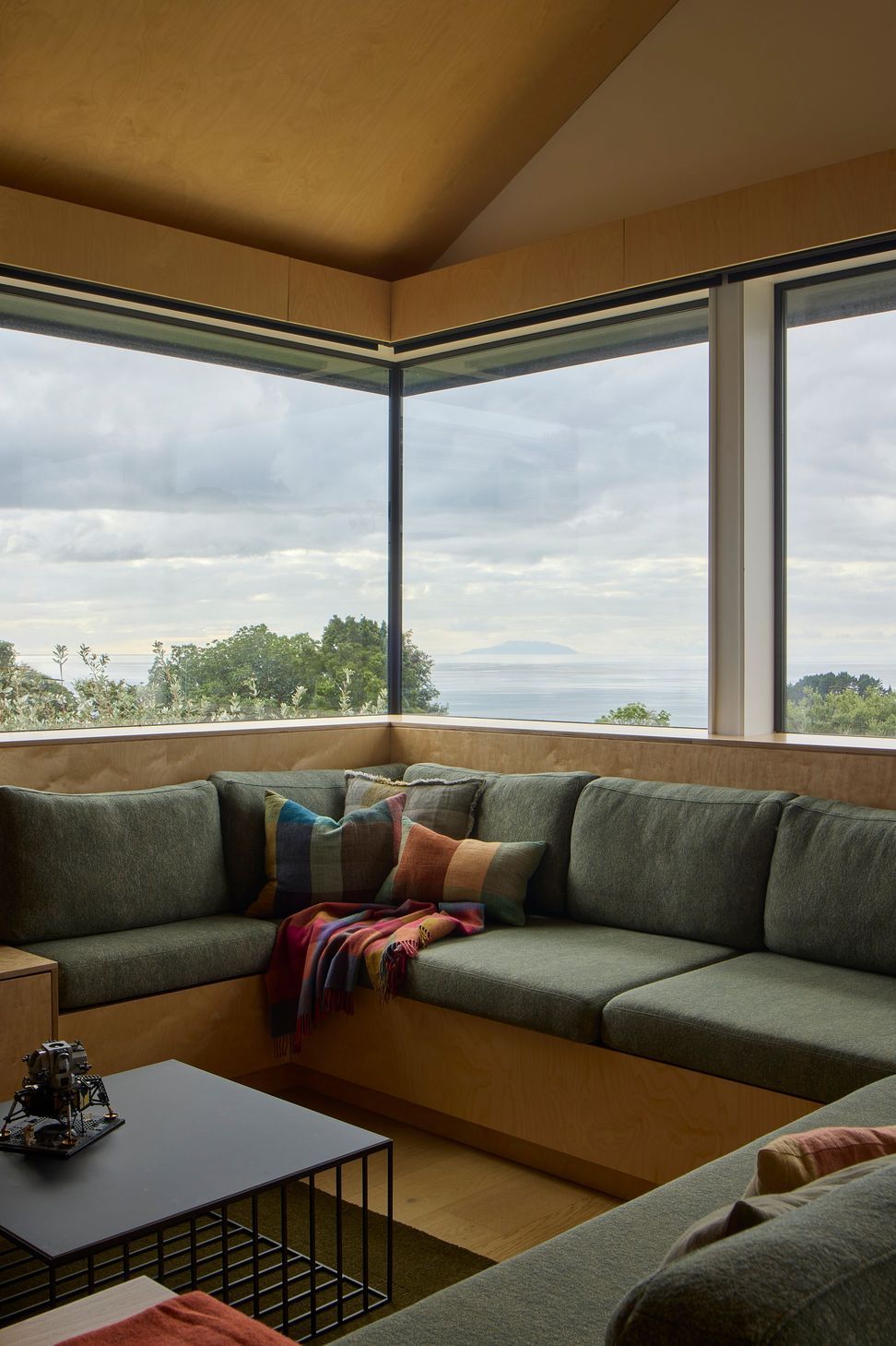 Built-in seating creates a comfortable gathering space within the living room. Upholstered in green fabric, it frames views across the landscape towards the ocean beyond.