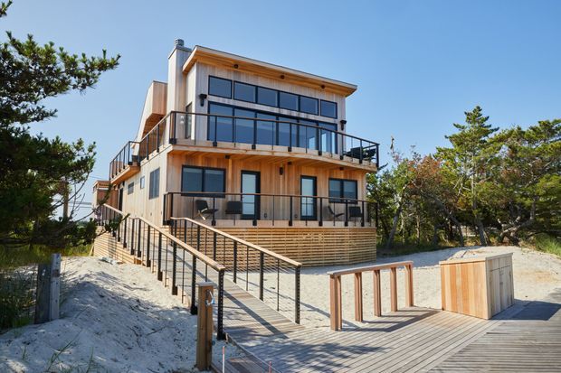 Contemporary multi-level coastal home set among sandy vegetation, with extensive timber cladding, dark-framed glazing, broad balconies, black metal balustrades, and a timber deck connected by stairs and ramps.