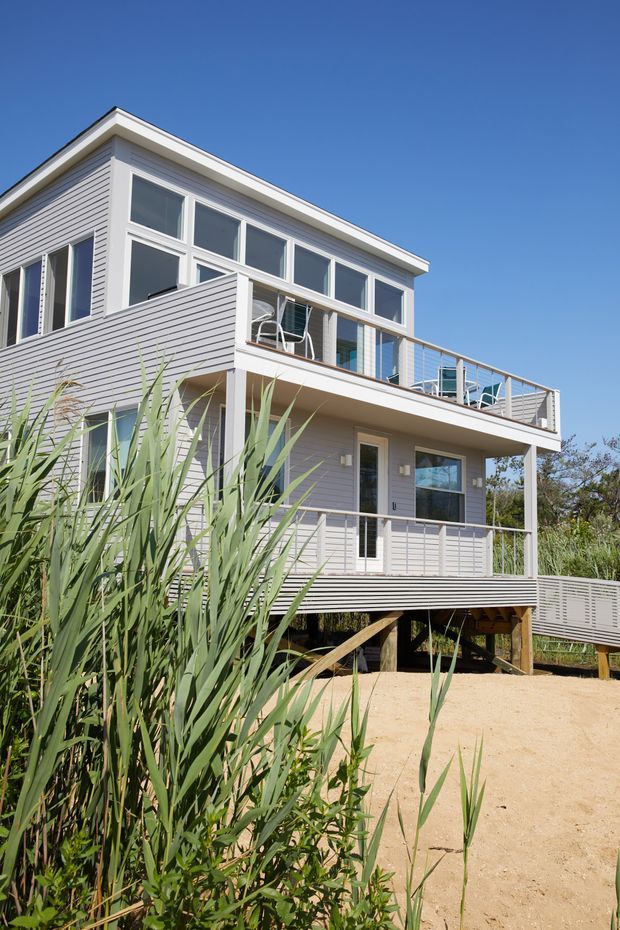 Exterior view of a raised, multi-level coastal-style house with pale horizontal weatherboard cladding, extensive white-framed glazing, and broad timber decks with simple metal balustrades. The building is elevated on posts above a sandy site and surrounded by tall coastal vegetation under a clear blue sky.