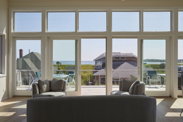 Bright living room viewed toward a broad wall of white-framed windows and glazed doors, opening to a balcony or terrace with outdoor seating. A dark sofa and upholstered armchairs face the view, while neighbouring coastal homes and a visible water horizon are seen outside. The room has a warm light timber floor and a high, airy glazed elevation.