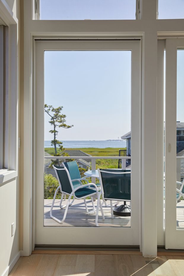 Bright interior view through a glazed exterior door onto a timber deck, with outdoor seating and a table overlooking a calm coastal waterfront. The room has pale walls, white-trimmed glazing and light timber flooring, while the deck and ocean landscape are visible beyond.