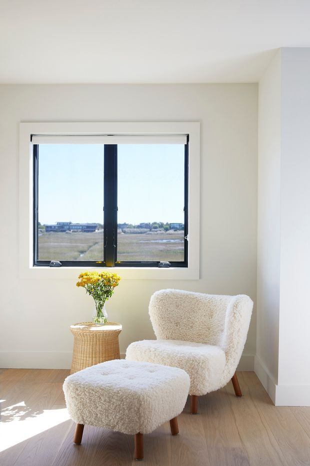 Bright, minimalist sitting corner with a cream boucle armchair and matching ottoman positioned beside a black-framed window. A small woven side table holds a vase of yellow flowers, while pale timber flooring and warm off-white walls create a calm, airy interior. The window overlooks an open landscape.